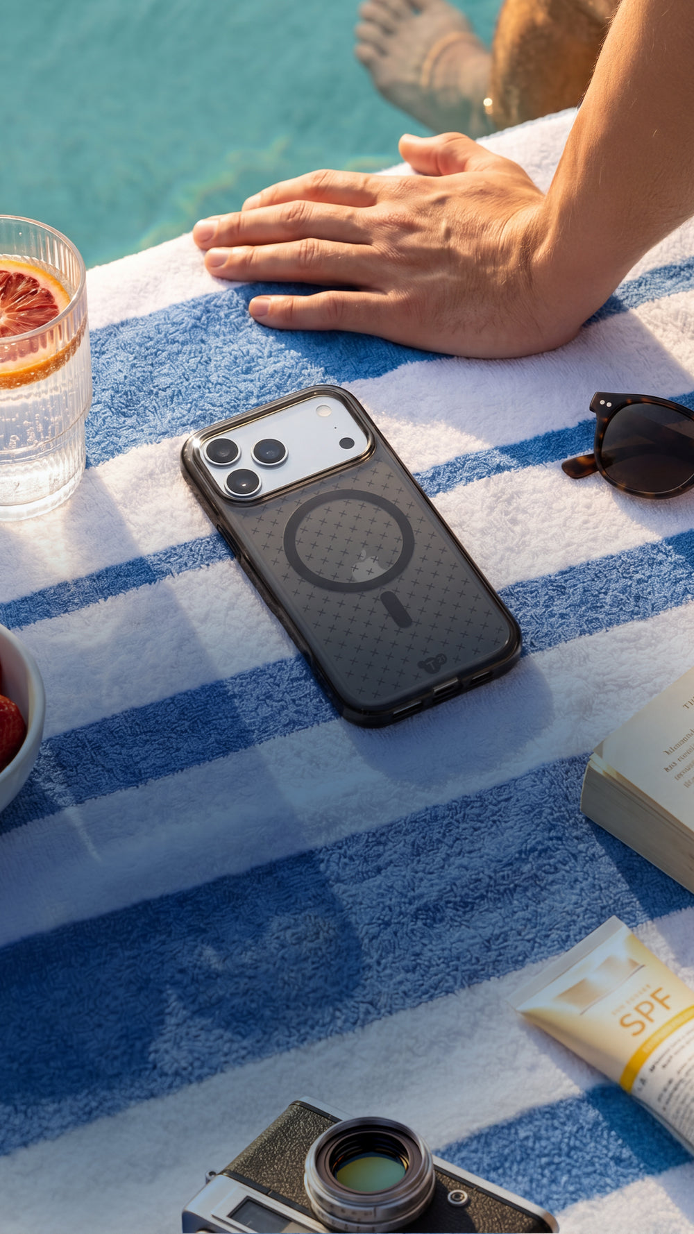 An iPhone in an EvoCheck case by Tech21 lays flat on a towel poolside next to some strawberries, a drink, sunglasses, and a camera.