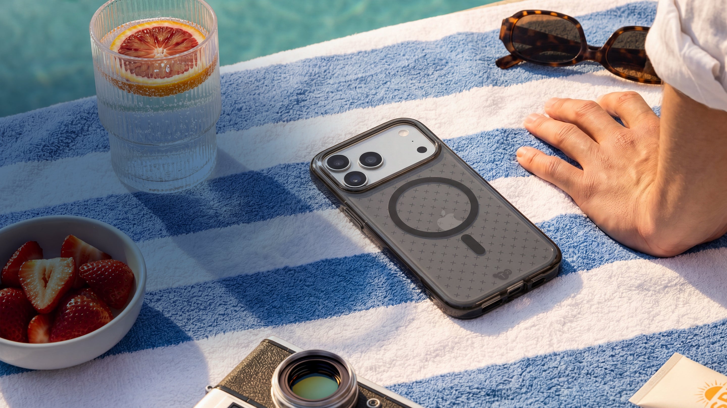 An iPhone in an EvoCheck case by Tech21 lays flat on a towel poolside next to some strawberries, a drink, sunglasses, and a camera.